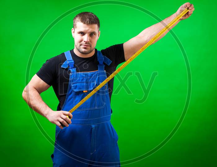 A Dark-Haired Male Builder In A Black T-Shirt And Blue Construction Jumpsuit Shows A Long Yellow Tape Rule On A Green Isolated Background