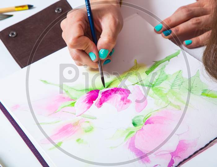 Close-Up Of The Artist Draws Watercolor In The Album For Drawing With A Thin Wooden Brush, On The Table A Pallet For Drawing And A Mug With Tools