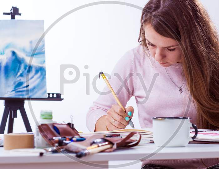 Young Dark-Haired Female Artist Paints A Picture Of A Watercolor Behind A White Table, In The Background A Picture Of The Sea In A Light Studio