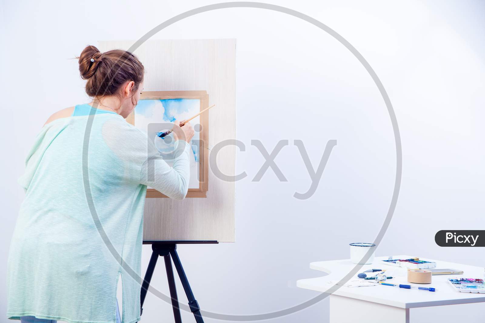 Young Dark-Haired Woman Artist In Blue Sweater Paints With Watercolor And Wooden Brush On White Paper Seascape With Single-Deck Yacht