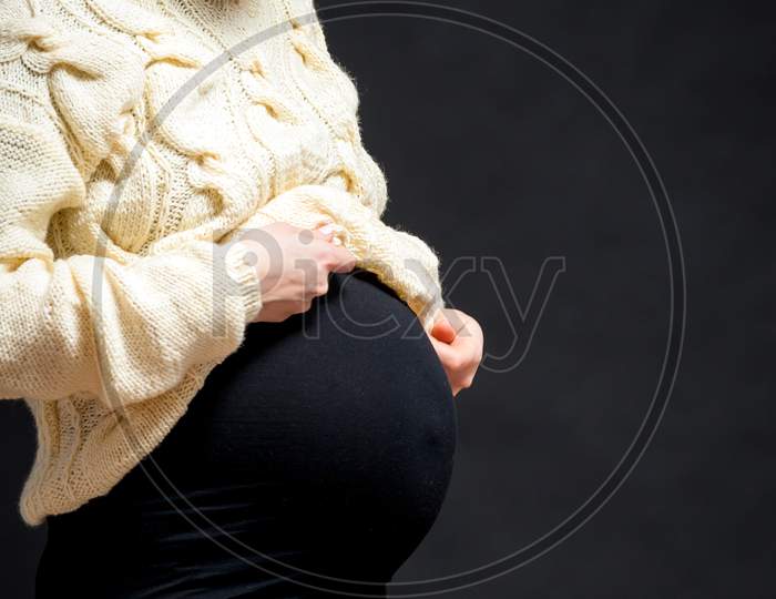 Close-Up Of A Young Pregnant Woman Woman In A Knitted Milk-Colored Sweater And A Black Dress Posing And Hugging Her Belly On A Black Isolated Background