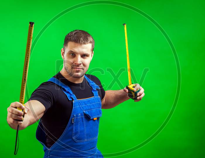 A Young Male Carpenter Wearing A Black T-Shirt And Blue Construction Jumpsuit Holds Two Long Yellow Tape Rule In His Hands For Construction Work On A Green Isolated Background.