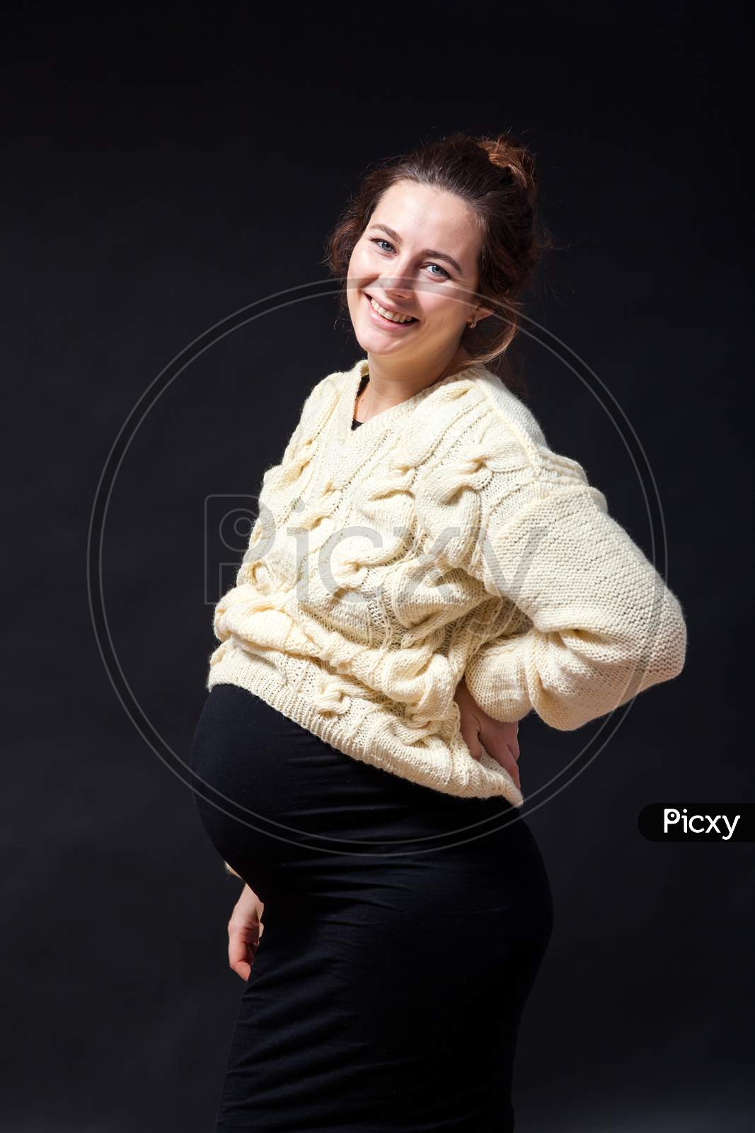 Close-Up Of A Young Pregnant Woman Woman In A Knitted Milk-Colored Sweater And A Black Dress Smiling And Holding Herself Behind Her Back On A Black Isolated Background