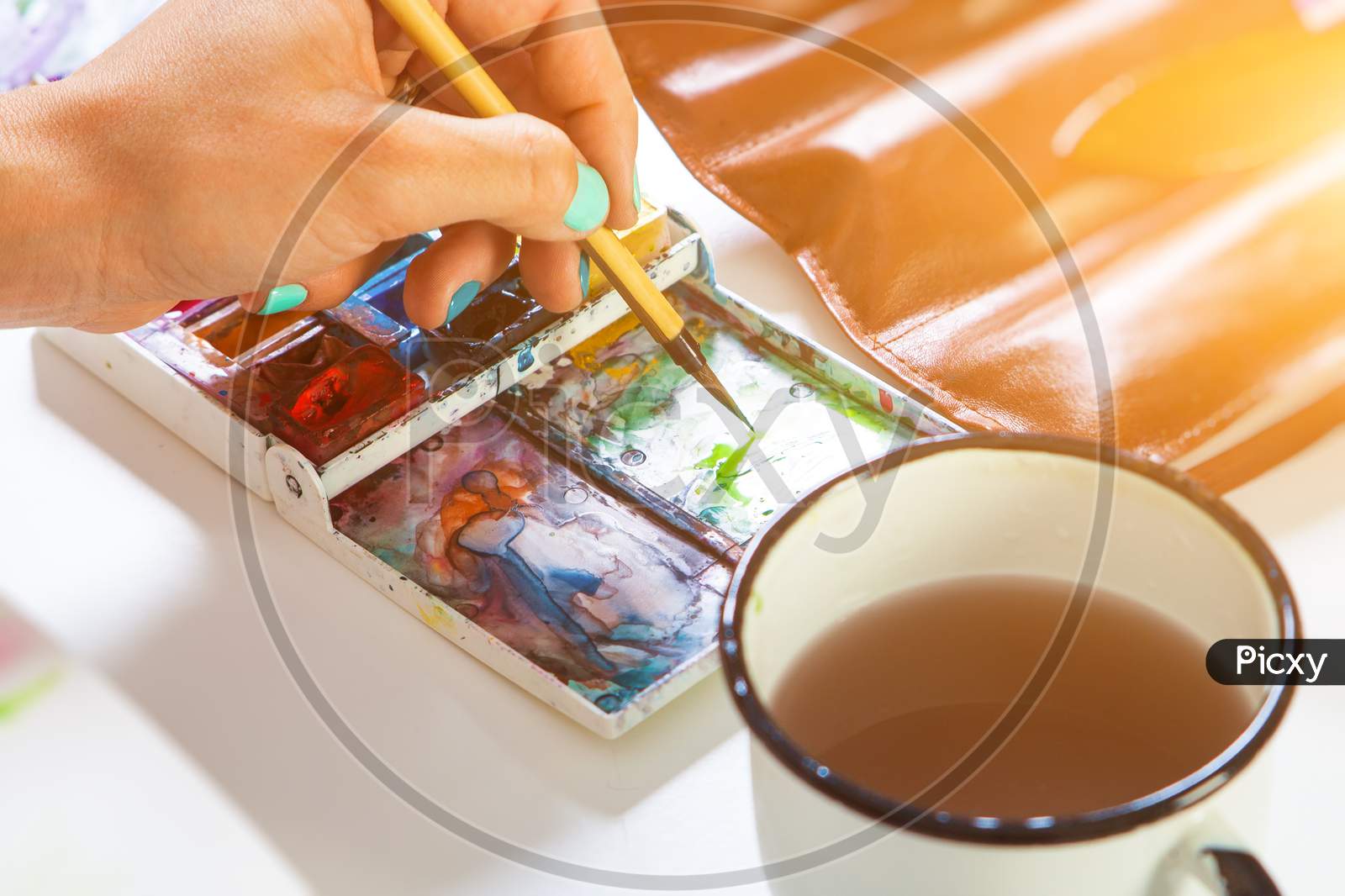 Close-Up Artist With Bright Nails Dabs A Brush In A Pallet With A Watercolor A Thin Wooden Brush, On The Table There Is A Leather Cover With Brushes And A Mug Of Water