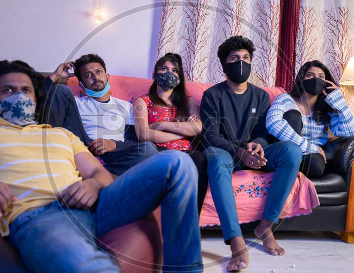 Group Of Friends With Medical Face Mask Busy Watching Movie Or Online Webs Series While Sitting On Sofa During College Holiday Due To Coronaviru Covid-19 Pandemic.