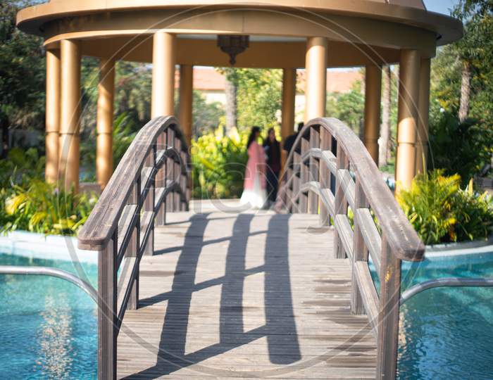 Locked Shot Of Arched Wooden Bridge With Swimming Pool On Both Sides With Blue Water And Out Of Focus People In The Distance Moving And Enjoying The Vacation In This Resort