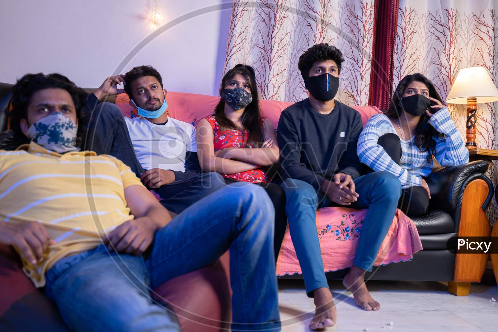 Group Of Friends With Medical Face Mask Busy Watching Movie Or Online Webs Series While Sitting On Sofa During College Holiday Due To Coronaviru Covid-19 Pandemic.