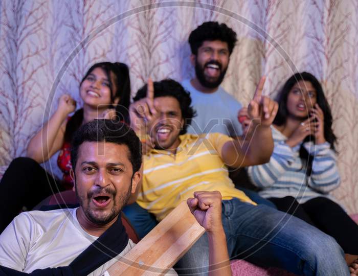 Group Of Young Sports Fans Holding Cricket Bat And Ball Screaming And Shouting For Sixer While Watching Cricket Match On Television From Home.