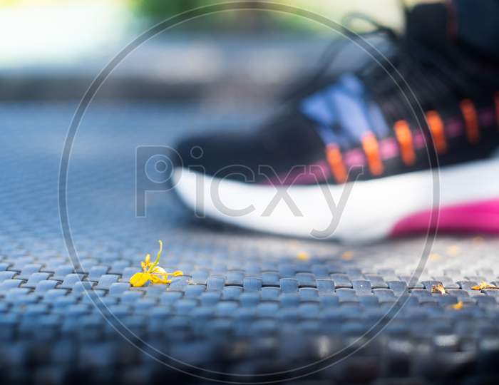 Macro Shot Showing Small Fallen Flower On A Wickerware Mat With A Colorful Sneaker Shoe Of A Strong Independent Powerful Woman Doing Yoga Exercise