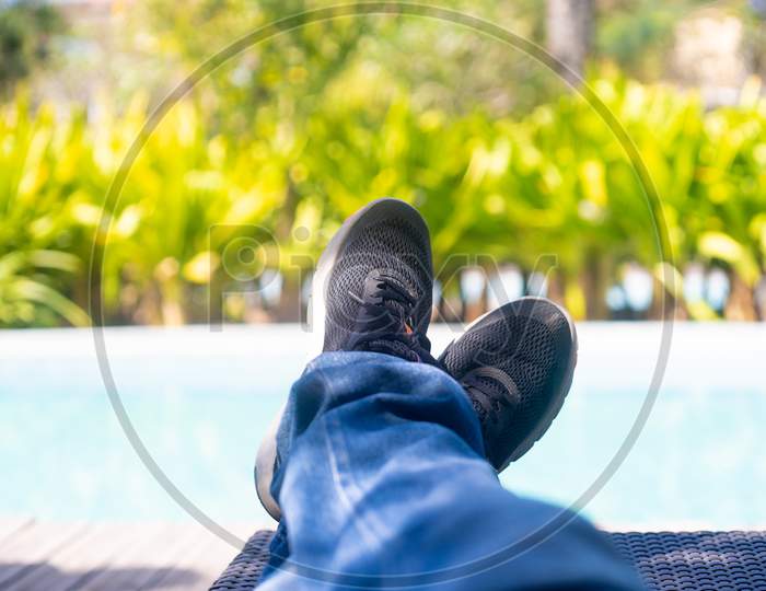 Man With Colorful Shoes And Blue Jeans Stretched Out On The Side Of A Pool With Plants Trees And Greenery As He Unwinds, Relaxes And Enjoys A Day In The Summer Sun