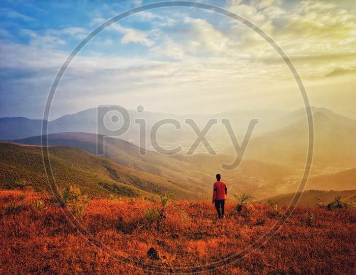 Young Man Standing On Top Of Cliff In Summer Mountains At Sunset And Enjoying View Of Nature