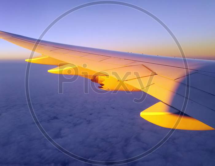 Wing Of Plane Over Dramatic White Fluffy Clouds. Airplane Flying On Blue Sky During Sunrise Or Sunset. Scenic View From Airplane Window. Commercial Airline Flight In The Morning With Sunlight
