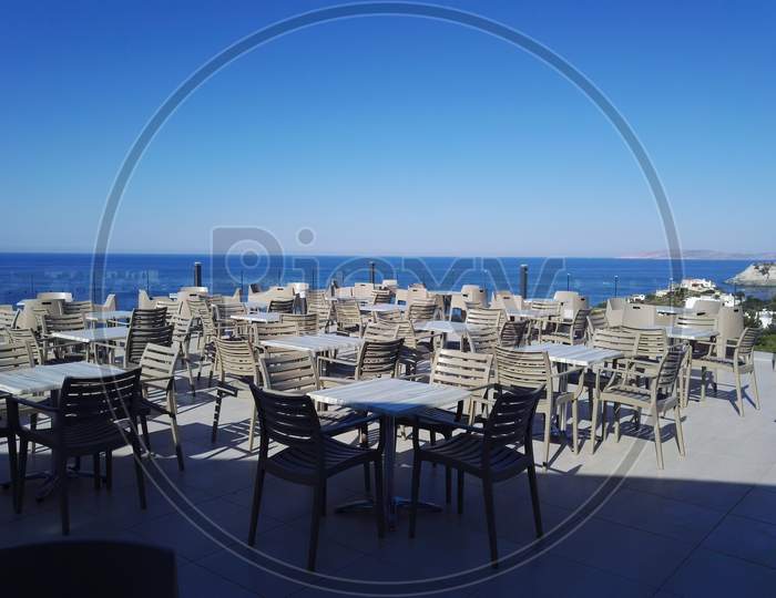 Santorini, Greece: The Setting Sunny Summer View Of A Big Terrace Roof With Cozy Tables And Chairs Showing Open Restaurant Against The Backdrop Of The Sea Ocean. Travel Invitation