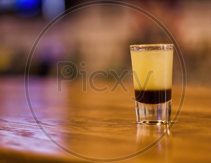 A Tequila Shot Of Cocktail Composed Of Different Syrup In Layers In A Small Glass On Wooden Table In A Bar. Art Of Alcohol Liquer Drink Making. Selective Focus With Blur Background.