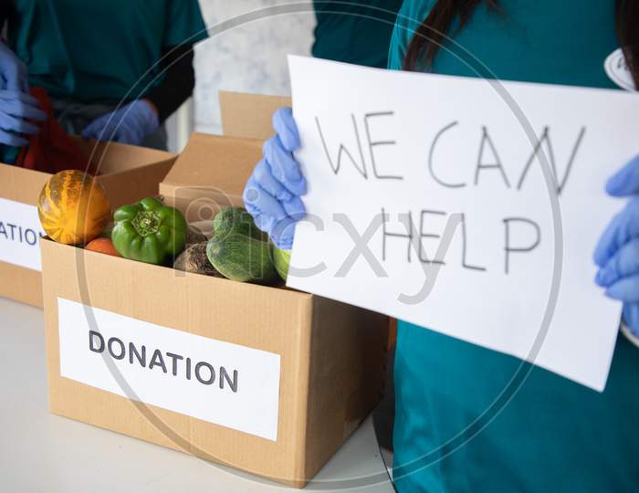 Focus On Vegetables, Volunteer Holding We Can Help Sign Board During Donations While Other Volunteers Busy Working Background - Concept Of Requesting For Donations.