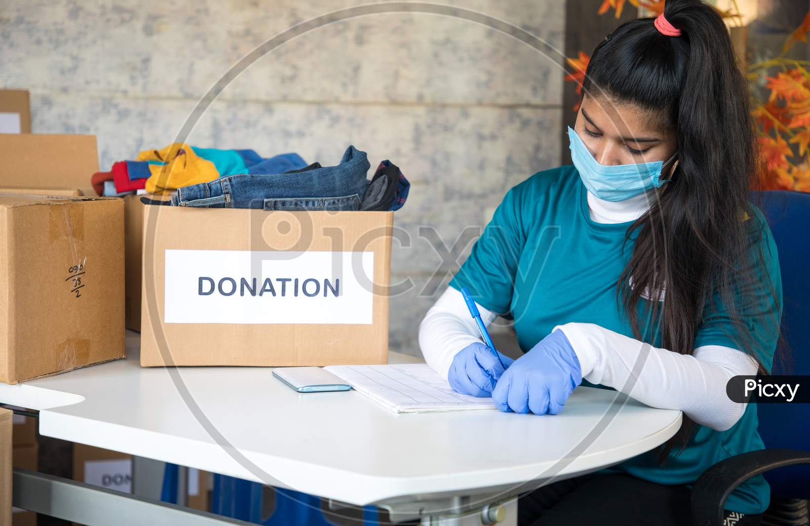 Young Girl Volunteer With Medical Face Mask And Gloves With Clipboard Noting Down Donors Information At Office During Coronavirus Covid-19 Pandemic.
