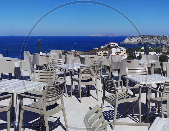 Panorama View Of Lots Of Traditional Wooden Tables And Chairs Of A Greek Tavern At Daytime And The View Of The Beach, Crete Island. Greece. Europe. The Invitation. The Dining Area Of Open Restaurant