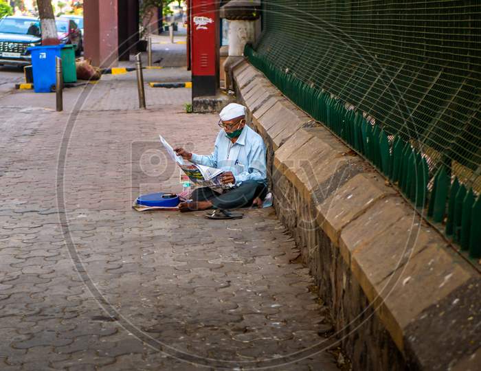 Old Man Wearing Glasses, Ethnic Maharashtrian Hat, Covid Mask And Reading Newspaper Seating On A Footpath Near Gateway Of India