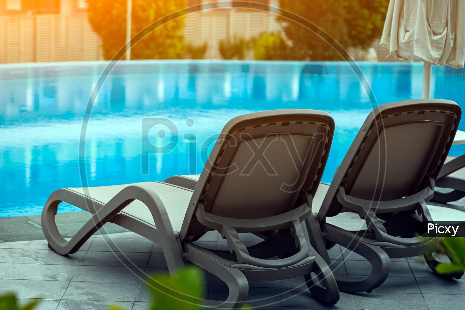 A Pair Of Comfortable Gray Sun Loungers Set Against The Backdrop Of A Beautiful Transparent Pool And Hotel. The Concept Of A Relaxing Seaside Vacation For Two