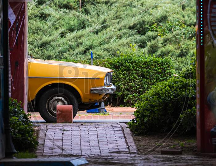 Yellow Old Car For Travel And Parked In A Narrow Small Street On A Summer Day Against The Backdrop Of A Garden With A Lot Of Bushes, Palms, Tropical Trees