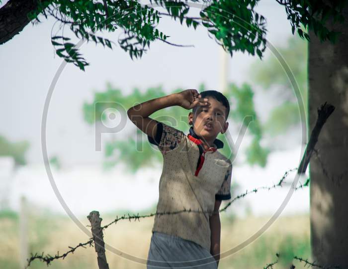Young Poor Village Boy Looking At The Camera Saluting Surprised Surrounded By Trees With A Barbed Wire Fence In Front Showing A Typical Village Scene