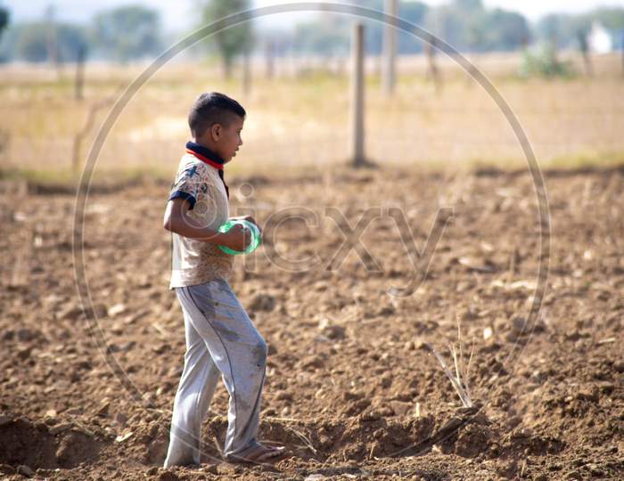 Young Indian Boy Carrying A Plastic Bottle Of Water To Father Working In The Field Sitting On Gunny Bag, Sacks Of Wheat Filled After Harvest In The Hot Summer Months In The Sun