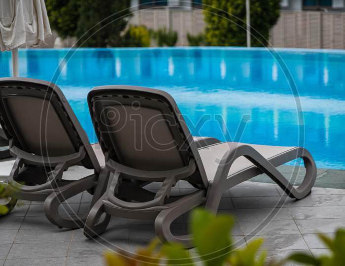 A Pair Of Comfortable Gray Sun Loungers Set Against The Backdrop Of A Beautiful Transparent Pool And Hotel. The Concept Of A Relaxing Seaside Vacation For Two