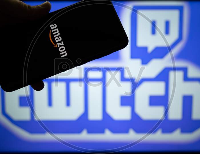 Man Holding Mobile Phone With Amazon Logo And Twitch Streaming Platform Name And Logo In The Background Showing This Platform With Full Time Earning Streamers