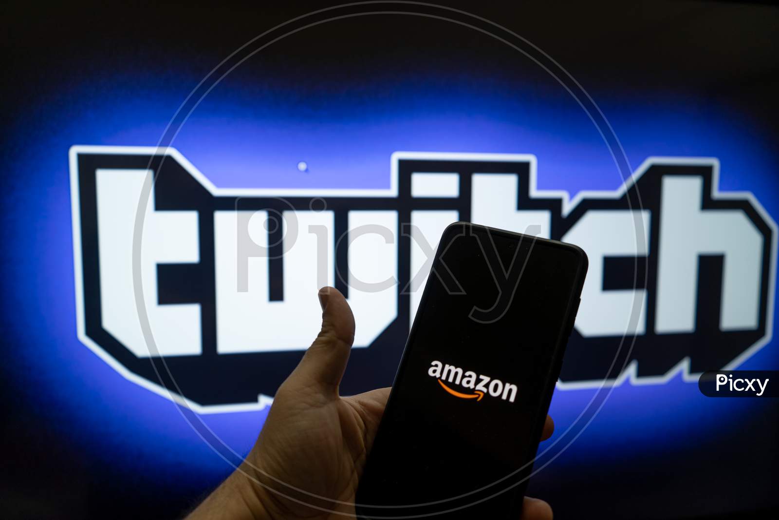 Man Holding Mobile Phone With Amazon Logo And Twitch Streaming Platform Name And Logo In The Background Showing This Platform With Full Time Earning Streamers