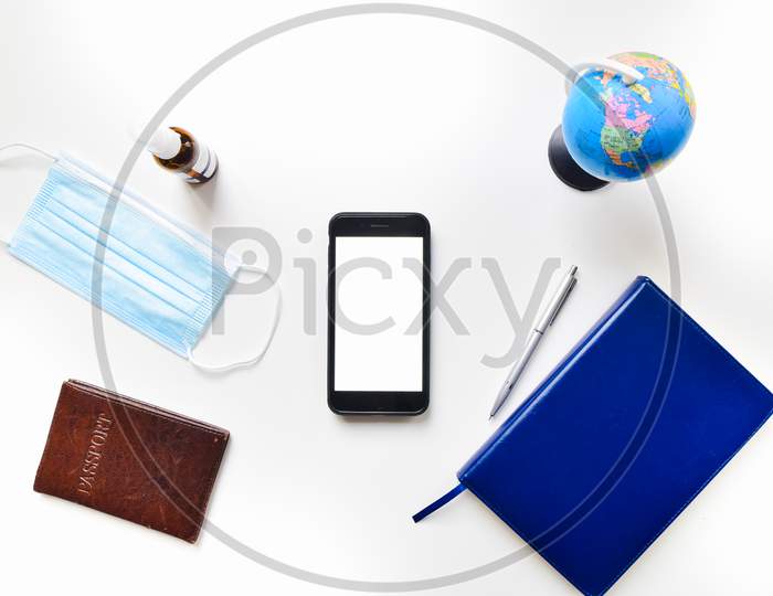 Top View Flat Lay Mobile Phone Green Screen Copypaste With Blue Notebook, Passport, Sanitizer, Blue Face Mask, Global Stand On White Table Isolated Background.