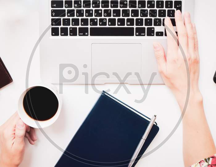White Office Desk Table With Blank Blue Notebook And Hand Holding Coffee Cup. Top View With Copy Space, Flat Lay Workspace Freelancer.