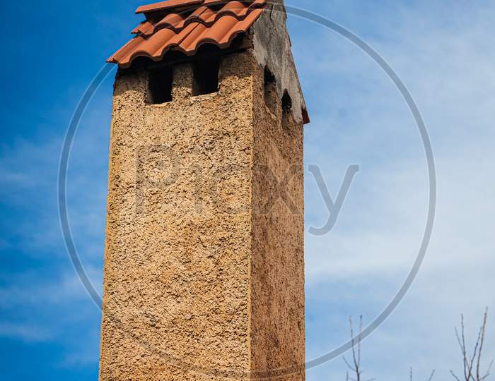 Stone Tall Chimney With Tiles On The Roof Of A Country House Against The Blue Sky
