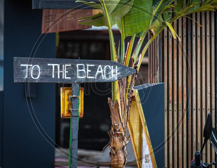 Wooden Signpost With The Inscription "To The Beach" On The Background Of A Tourist Street With A Cafe And A Palm Tree