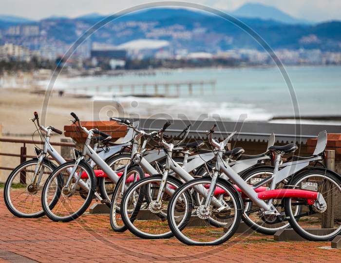 A Range Of Different Bicycles For Rent Parked Against The Backdrop Of The Beach And Sea