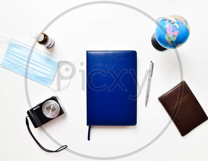 Flat Lay With Copy Space, Working From Home Concept During The Covid-19 Pandemic. A Smart Phone, Sunglasses, Laptop, Shoes, Passport, A Face Mask And Notepad Arranged On White Table Background