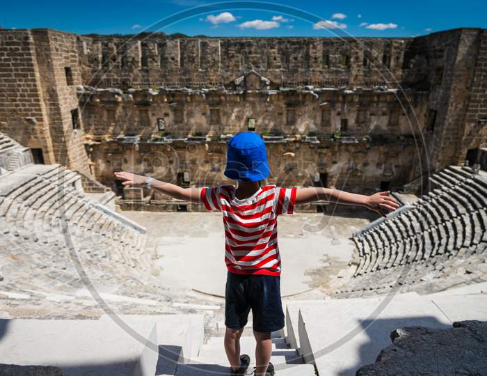 A Little Boy Tourist Spread His Arms Out To The Sides Against The Backdrop Of The Roman Amphitheater Of The Ancient City Of Aspendos Near Antalya, Turkey. Ancient Ruined City