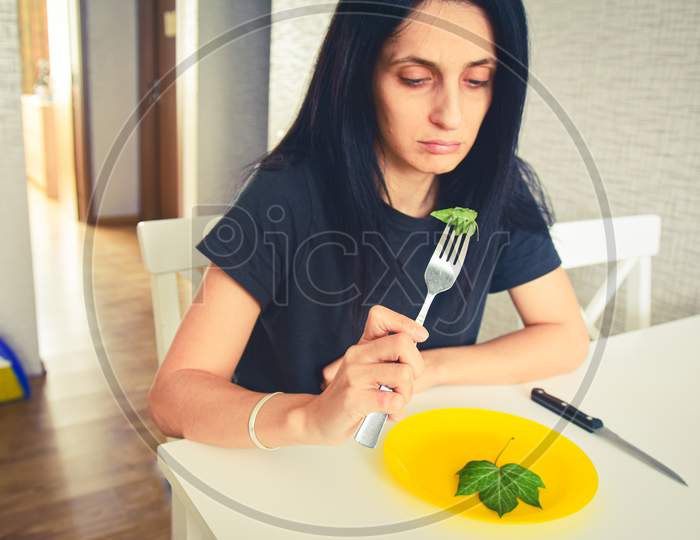 Young Attractive Female Eat Green Leafs Unhappy In Kitchen To Loose Weight. Concept Unbalanced Diet And Unhealthy Raw Foods