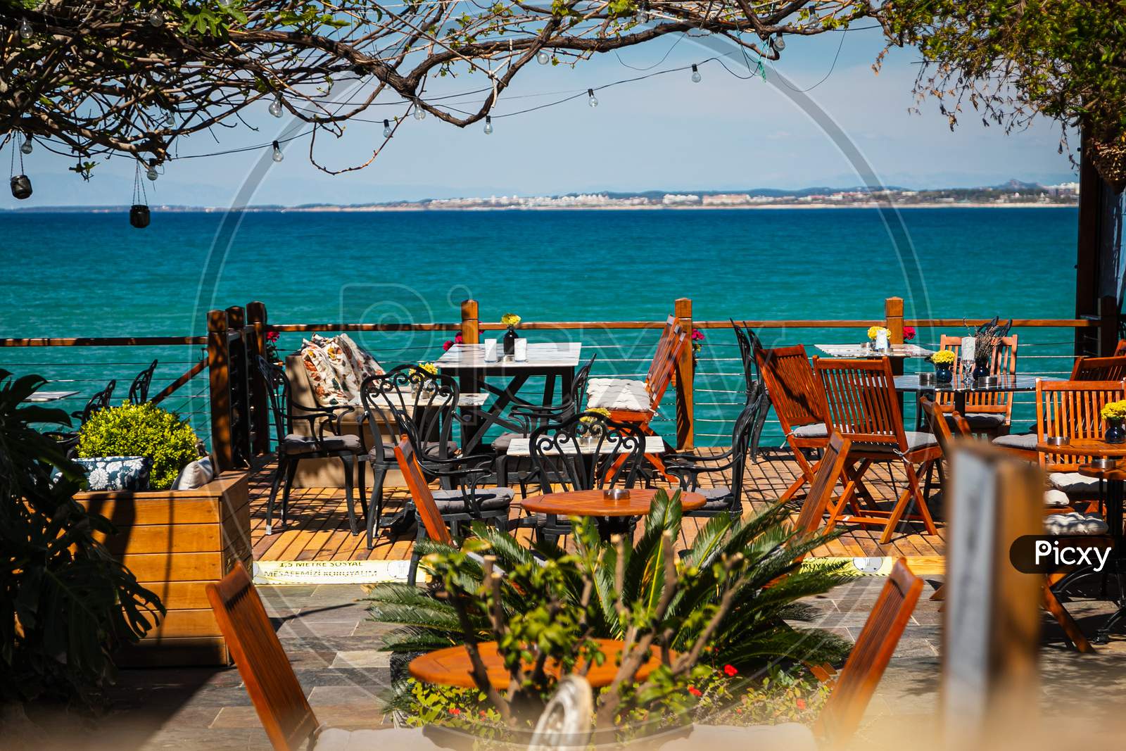 The Concept Of A Measured Rest On The Seashore. The Perfect Place For A Date. Restaurant With Wooden Furniture, Lanterns, Palm Trees And Vegetation On The Seaside Of The Ancient City