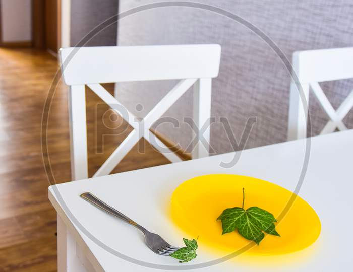Kitchen White Table With With Wild Green Leaf On Yellow Plate. Extreme Diet And Slimming Concept.