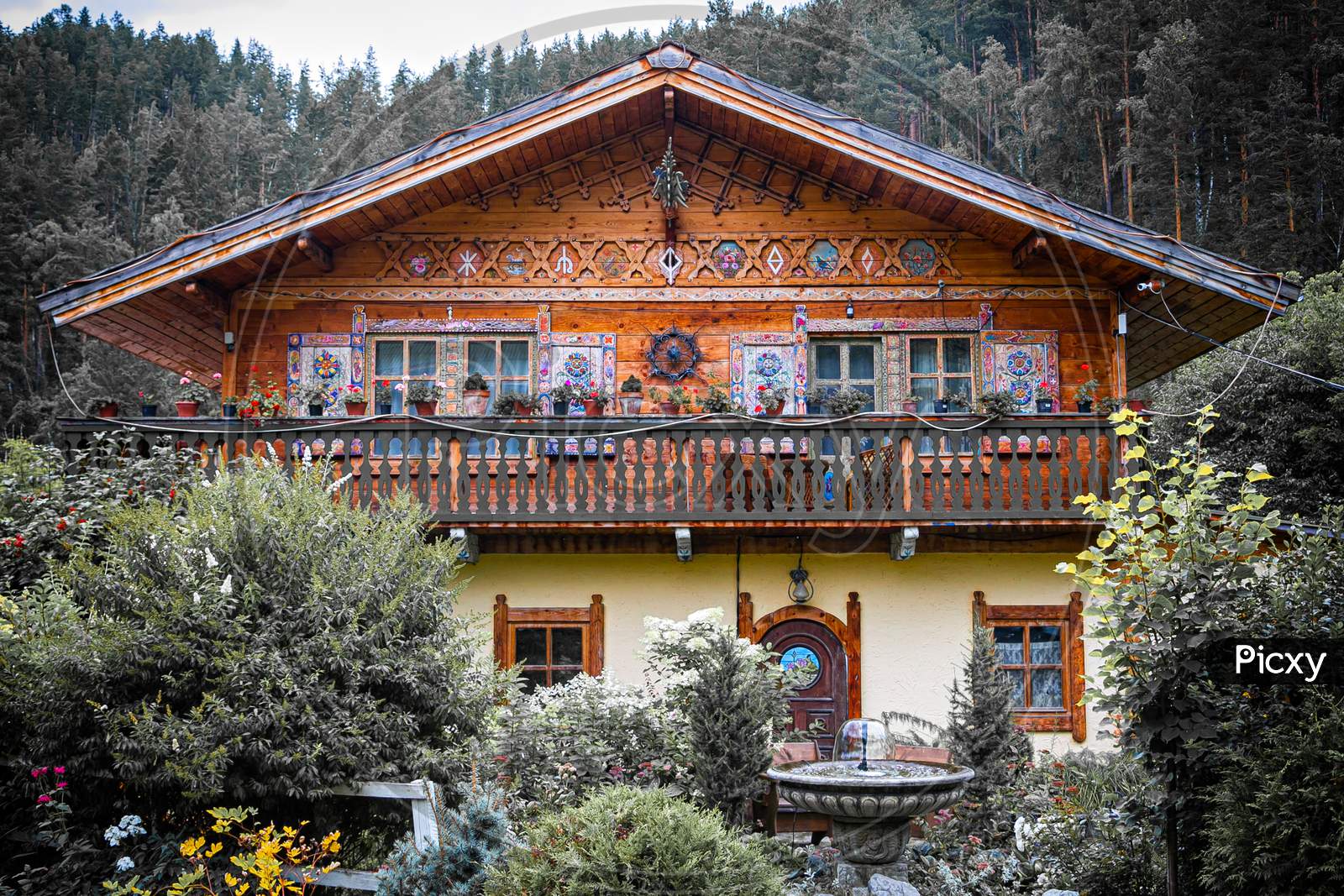 Authentic Wooden Two-Story House With A Fountain With Painted Walls, Shutters And Windows Stands, Surrounded By A Lot Of Trees, Bushes And Various Flowers And Plants, In The Background Mountains And Forest