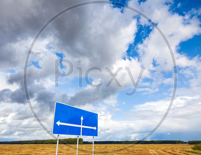 Summer Landscape: A Wheat Field With A Sign Indicating Directions And Without Text On The Background Of Trees And A Stunningly Blue Sky With Lots Of Clouds On A Bright Sunny Day. Space For Text On A Road Sign