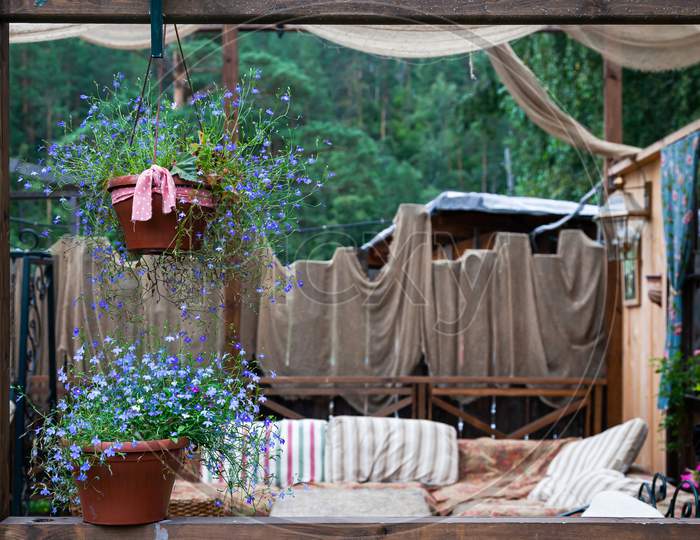 Beautiful Scenery Of The Rustic Veranda: Pots With Purple Flowers, A Lamp, Decoration With Various Fabrics And A Sofa