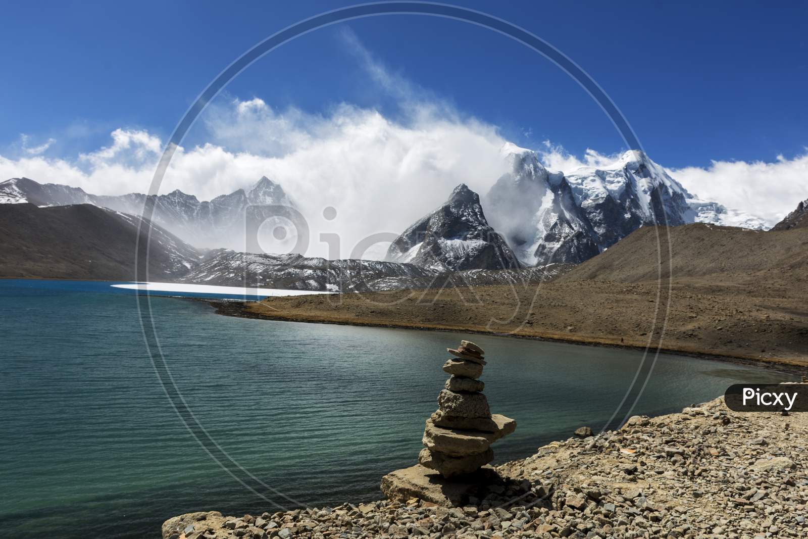 A Beautiful Landscape Of Gurudongmar Lake With Rocks In Forte Ground And Mountain With Blue Sky In Background. Gurudongmar Lake One Of The Highest Lake Of The World And India Located At An Altitude Of 17,800 Ft, In The Indian State Of Sikkim