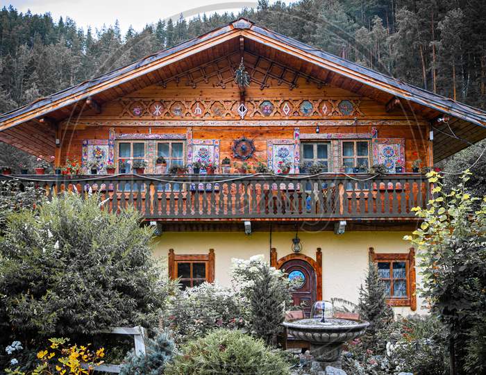 Authentic Wooden Two-Story House With A Fountain With Painted Walls, Shutters And Windows Stands, Surrounded By A Lot Of Trees, Bushes And Various Flowers And Plants, In The Background Mountains And Forest