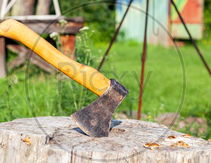 Close-Up Of A Large Ax With A Wooden Handle Stuck In A Wooden Stump After Chopping Firewood On A Green Background On A Warm Summer Day