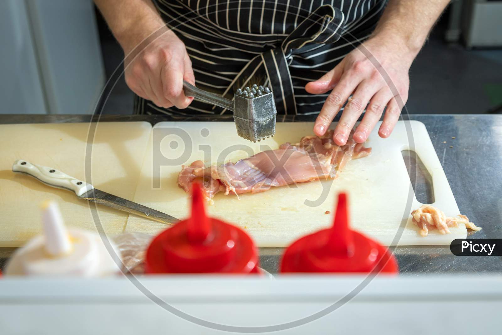 Close Up Male Cook In An Apron With A Metal Hammer Beats Chicken Meat For Softness On A Cutting Board. Cooking Process