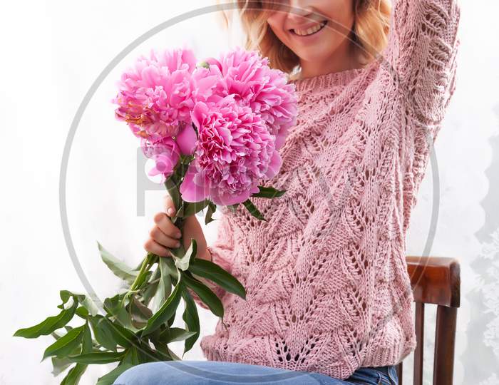 A Young Woman In Delicate Jeans And A Knitted Sweater Poses And Holds In Her Hands A Beautiful Huge Bouquet Of Peonies In The Morning In The Bedroom. Romantic Portrait Of A Girl On A Gentle Background