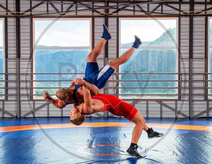 Two Men In Sports Wrestling Tights And Wrestling During A Traditional Greco-Roman Wrestling In Fight On A Wrestling Mat Against The Backdrop Of Mountains. Wrestler Throws His Opponent’S Chest Through