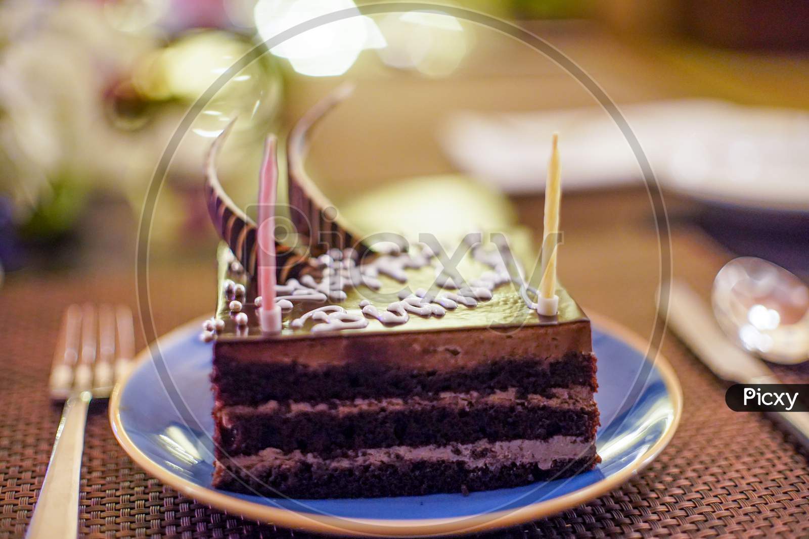 Shot Of Cake With Sparkling Candles With Flames And Sparks Flying Out With An Out Of Focus Background Of Flowers, Lights Showing A Birthday Celebration, Wedding Or An Anniversary