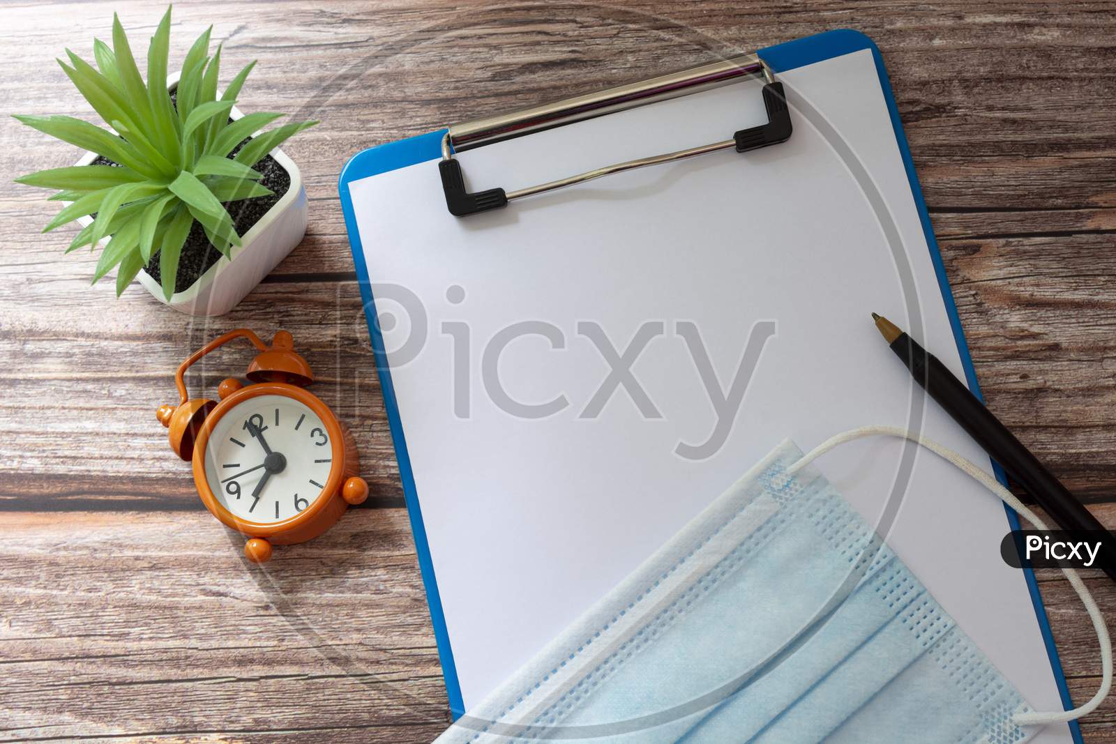 Face Mask On A Clipboard With Pen, Alarm Clock And Potted Plant On Wooden Desk. For Text Use